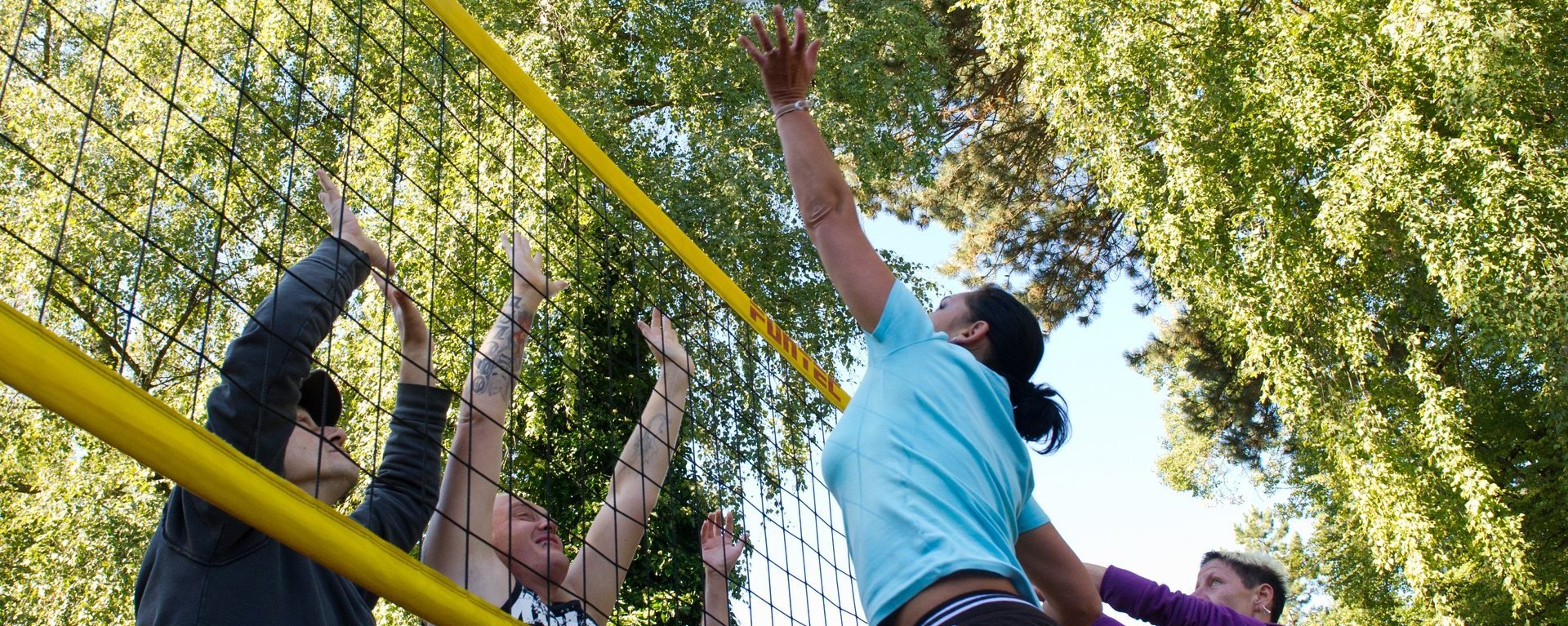 Action: eine Gruppe Frauen und Männer spielen Volleyball - eine Frau "lupft" gerade den Ball über den "Block"
