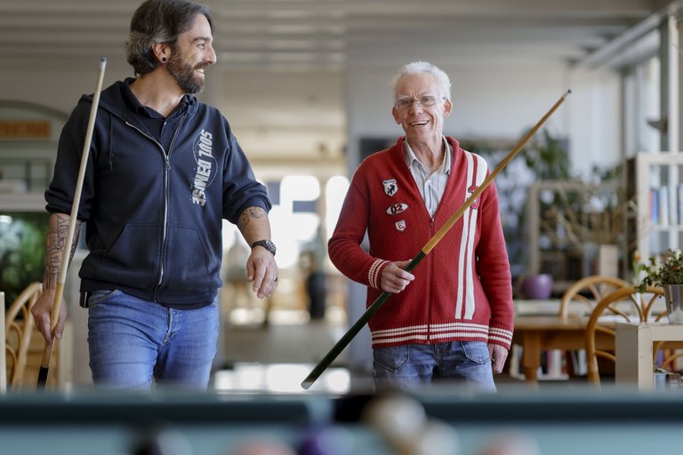 Zwei Männer laufen mit zwei Billardqueues in den Händen auf den Billardtisch zu.