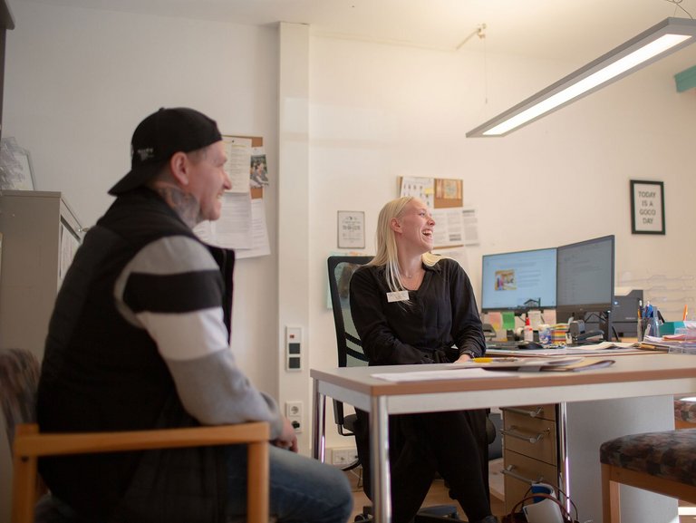 Eine freundliche Mitarbeiterin im Büro und ein Mann mit Base-Cap lachen herzhaft