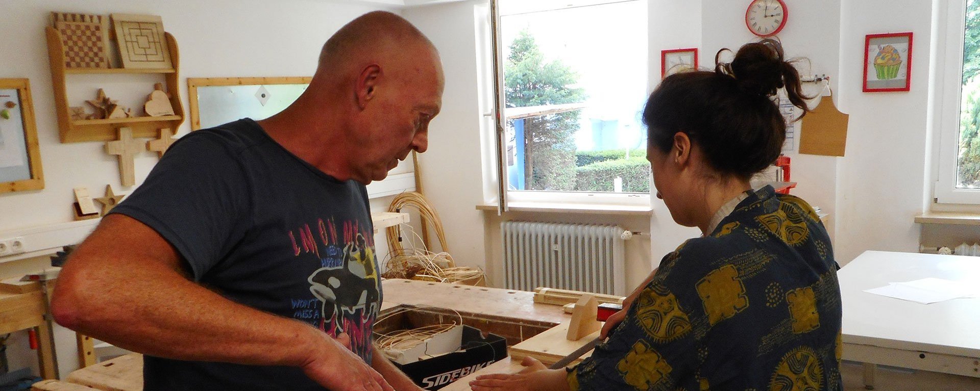 Werkstatt: ein Mann hilft einer Frau beim Holz-Raspeln an einer Werkbank