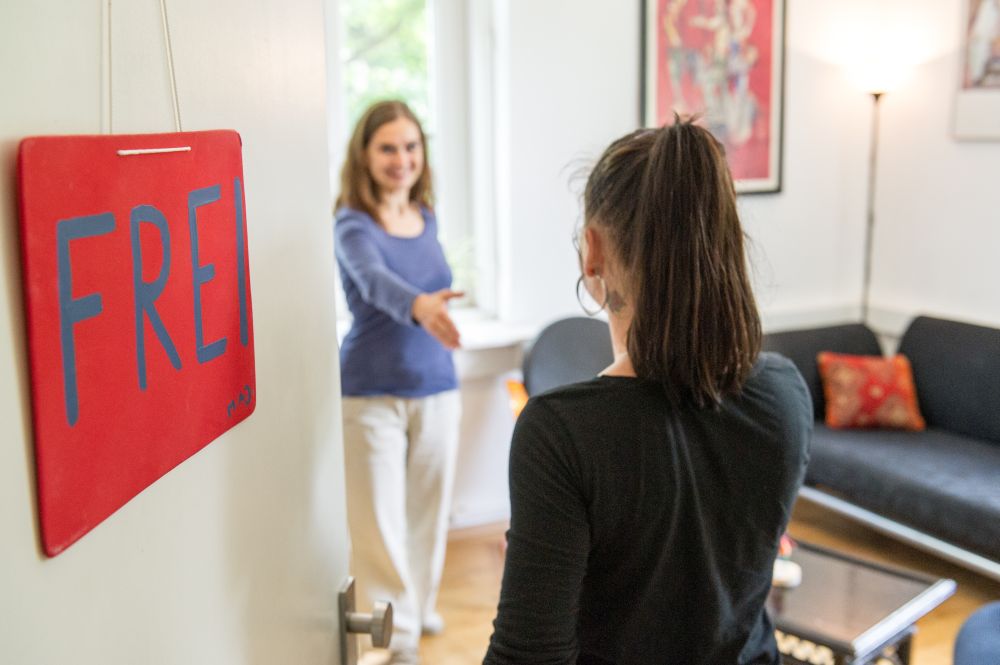 Eingangstüre mit einem, in rot, gemalten Schild und der Aufschrift: "FREI". Im Hintergrund begrüßen sich zwei Frauen